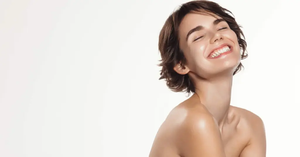 A smiling woman with short brown hair and bare shoulders tilts her head up with eyes closed against a white background for Peptide Therapy in Suwanee, GA.